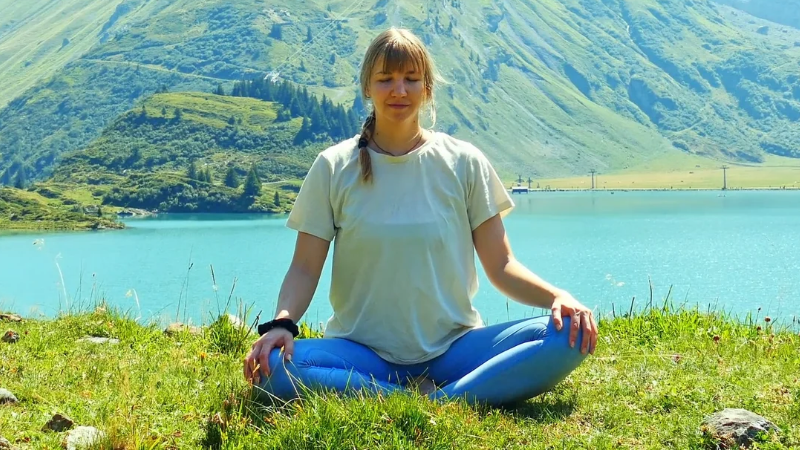 Yogalehrerin sitzt im Schneidersitz mit geschlossenen Augen auf einer Bergwiese vor einem türkisfarbenen See