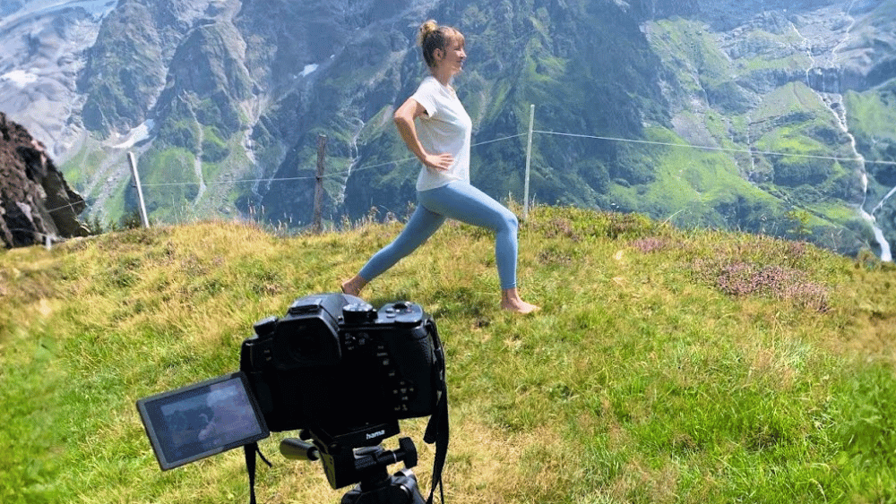 Kamera auf Stativ filmt Tamara von Office Relief bei einer Yoga-Übung im Ausfallschritt auf einer Bergwiese, dahinter Felswände und ein Wasserfall